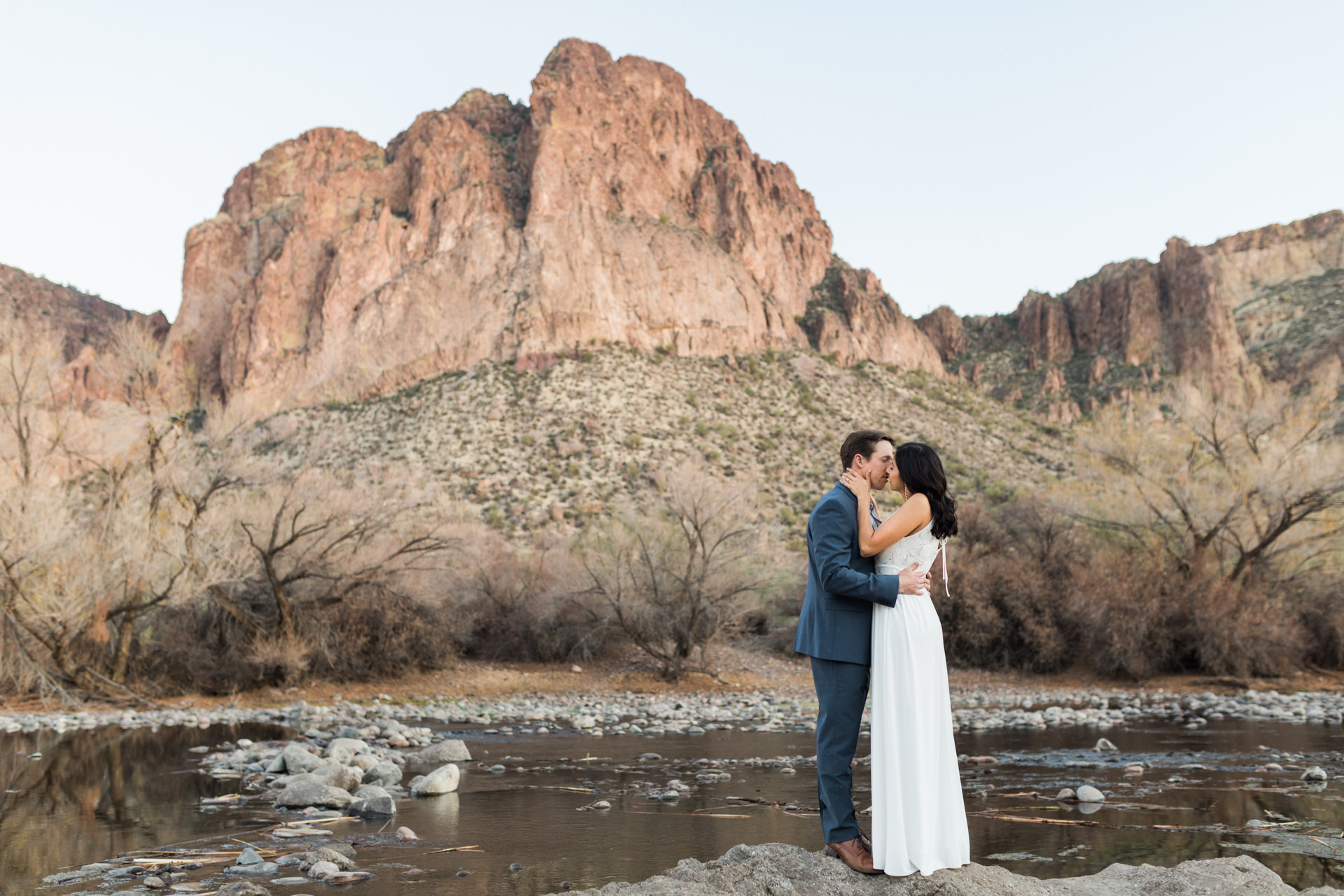 bride and groom kissing at river with mountain in backdrop